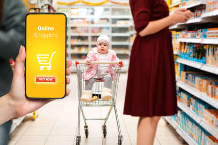 Shopping. Cute baby sitting in a shopping cart in the supermarket, next to his mom. The hand on the left side holds a smartphone. The concept of online shopping.の写真素材