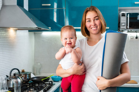Home yoga. A young smiling mother holds her baby in her arms, and a sports mat rolled up. In the background is the kitchen. The concept of sports activity at home.の写真素材