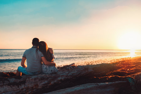 Summer vacation. A young couple posing against the sea, sitting on the rocks in an embrace. Rear view. Copy space. Sunset.の写真素材