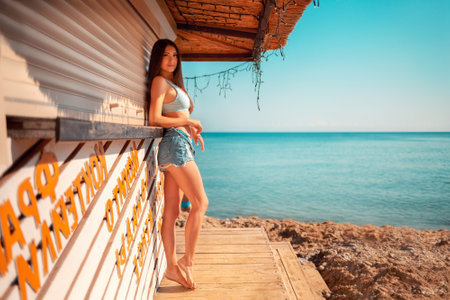 Beautiful tanned woman posing near a closed beach bar. Sea and sky in the background. Side view. Summer vacation and fashion concept.の写真素材