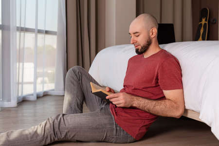 Portrait of a young, bearded, bald man is sitting on the floor at home reading a book. The concept...
