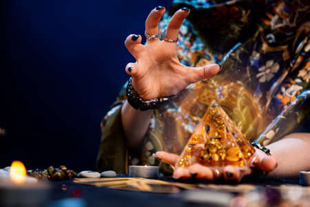 The fortune teller holds a glass pyramid with gold in her hands and casts a spell over it. Magic aura. Close-up. Copy space. The concept of divination, magic and esotericism.の写真素材