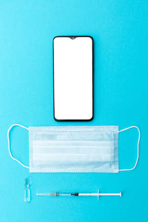 Cellphone with empty screen, vaccine, protective mask and syringe on a blue background. Flat lay. Mock up. Vertical. Concept of vaccination.の写真素材