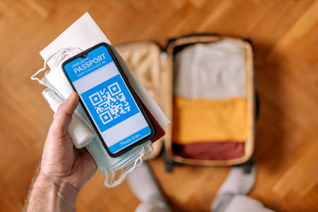 New normal concept. A male's hand hold a protective mask, antiseptic and smartphone with an immune digital health passport. In the background, a suitcase with clothes in a blur. Flat lay.の写真素材