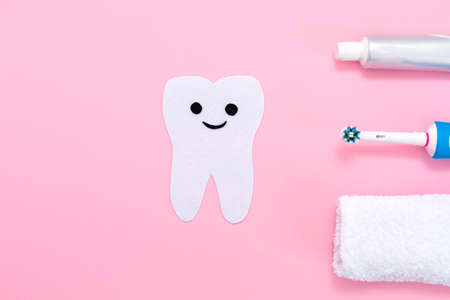 A silhouette of a tooth cut out of felt with a smiling cartoon face, next to an electric toothbrush, towel and toothpaste. Pink background. Hygiene of the oral cavity.の写真素材