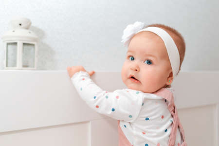 Portrait of a baby girl with a headband on her head, holding her hands to the crib. Concept of...