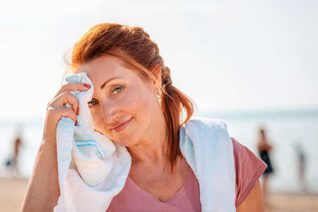 Portrait of an adult woman in sportswear smiling and wiping the sweat from her forehead with a towel. In the background, the sea and the beach. The concept of activity and communication.の写真素材