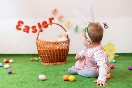 A little girl with rabbit ears, posing on an artificial lawn looking at the background with a basket and eggs. Rear view. Easter concept.の写真素材