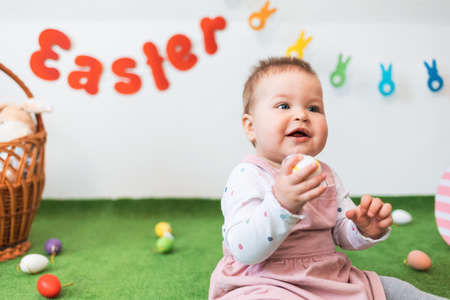 Portrait of a little happy Caucasian girl in a dress, sitting on a green rug with an Easter egg in her hand. In the background, Easter decorations.の写真素材