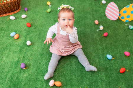 Little pretty baby girl in a dress and a floral band, sitting on a green rug with decorative eggs and basket. Easter holiday. Top view.の写真素材