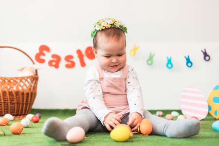 Portrait of a small happy Caucasian girl in a dress and a floral band, sitting on a green rug with an Easter egg in her hands. In the background, Easter decorations and a basket.の写真素材
