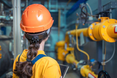 A female engineer in uniform and a protective helmet, holding a tablet and conducting an inspection of the equipment. The view from the back. The concept of industrial production.の写真素材