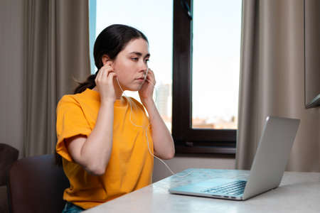 Portrait of a young pretty woman sits at a desk and puts on headphones, studying on a laptop. A...
