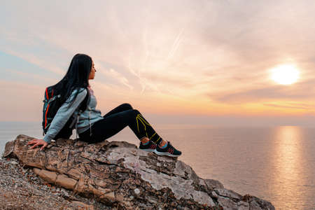 The concept of Hiking and sports recreation. A woman with a backpack on her back sits on a rock in...
