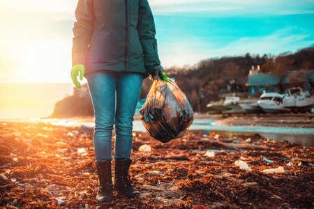 Day of Earth. A female volunteer stands on a muddy beach with a bag of garbage. Sunset.の写真素材