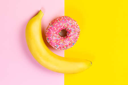 The concept of choice and nutrition. Banana and donut with pink icing on a yellow-pink background. Top view.の写真素材
