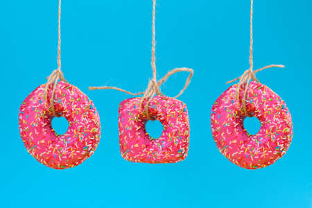 Three donuts of different shapes with pink frosting, suspended on a string. Close-up. Blue background.の写真素材