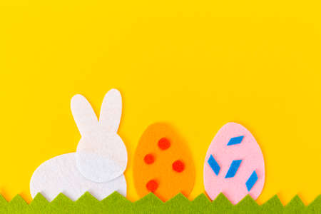 Cut out of felt applications of two colorful eggs and silhouette white rabbit on the grass. Yellow background. Flat lay. Easter holiday. Copy space.の写真素材