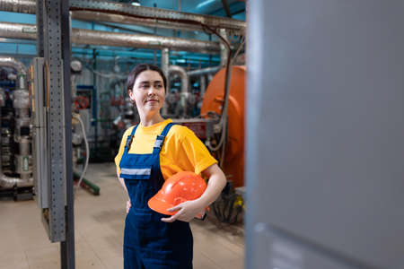 The concept of industrial manufactory. A young smiling woman in uniform holds a safety helmet in her hand. Boiler room on the background.の写真素材