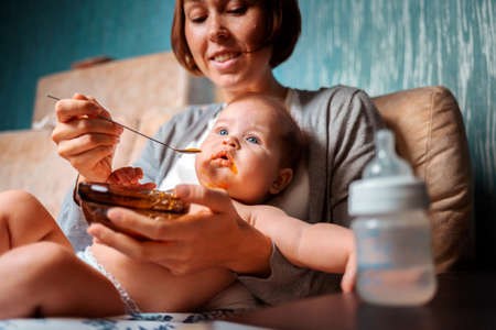 Mother is sitting in a armchair and holding a baby in her arms, who is eating from a bottle. Side view. Concept of infant feeding and weaning.の写真素材