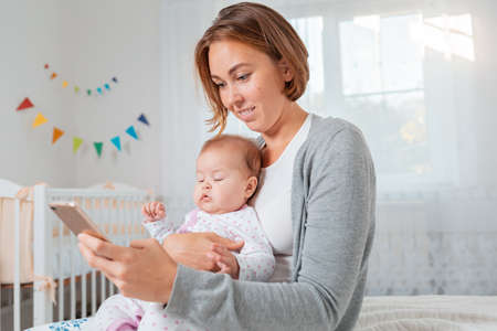 A smiling mother holds a sleeping child in her arms and using her smartphone. Childrens room on the...