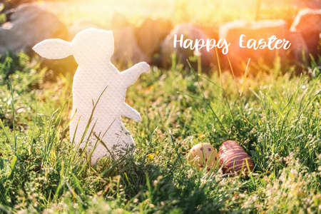 The concept of Easter holidays. Decorative gift handmade product in the form of a rabbit, located next to the painted eggs. Green grass in the background. Light and inscription "Happy ears".の写真素材