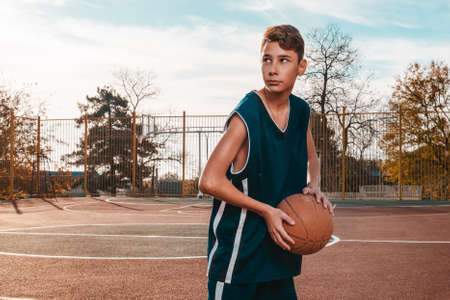 Sports and basketball. A young teenager in a blue tracksuit poses with a basketball in his hands and looks away. In the background, a basketball court. Copy space. Horizontal.の写真素材