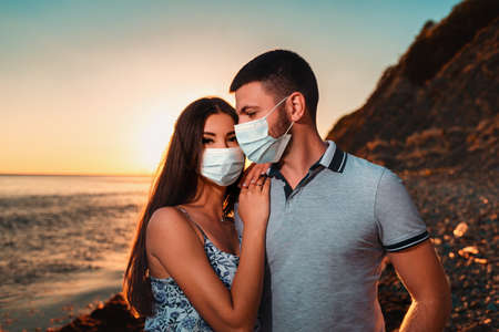 A young couple in protective masks posing on the sunset of the sea. In the background, the sea. The concept of travel during the coronavirus pandemic.の写真素材