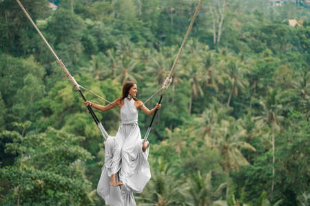 Tanned beautiful woman in a long white dress with a train, riding on a swing. In the background, a rainforest and palm trees. Copy space.の写真素材