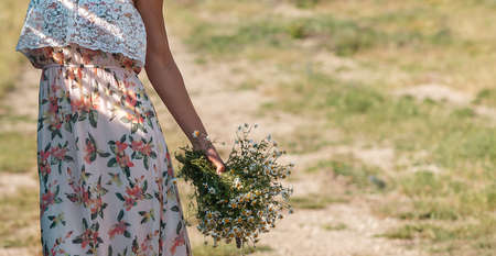 Summer time. A woman in a dress, holding a lush bouquet of daisies. In the background, a rural road overgrown with grass. Close up.の写真素材
