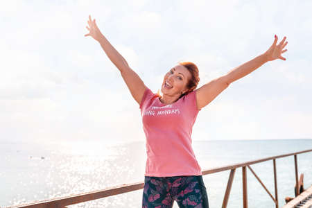 An adult woman in sportswear smiles happily and poses with her hands raised in the air against the...