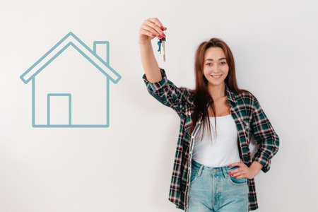 Buying and renting real estate. A smiling woman in a plaid shirt holds a bunch of keys, and a turquoise house is painted on the wall. Hand in focus. White background.の写真素材