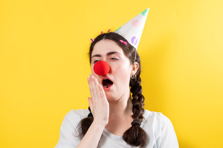 April's fool day concept. Portrait of a bored, yawning woman in a cap and with a red round false nose. Yellow background.の写真素材