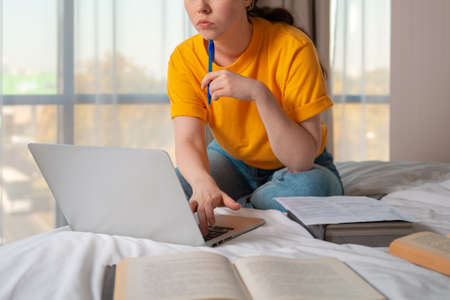 Remote work. A Caucasian woman is sitting on a bed and working on a laptop. Close up. Books are lying next to each other. The concept of online education.の写真素材