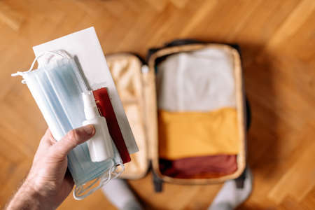 Male's hand hold antiseptic, masks and passport. In the background, in a blur, lies an open suitcase with clothes on top. The concept of a new normal and travel during covid-19 pandemic.の写真素材