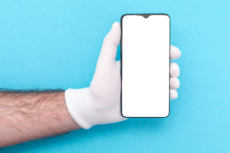 A male hand in white gloves holds a smartphone with a white screen. Mock up. Blue background. Copy space. Business and advertising concepts.の写真素材