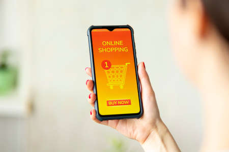 A woman holds a cellphone in her hand. An app with shopping cart is opened on the device screen. Blurred background. The concept of online shopping and consumerism.の写真素材