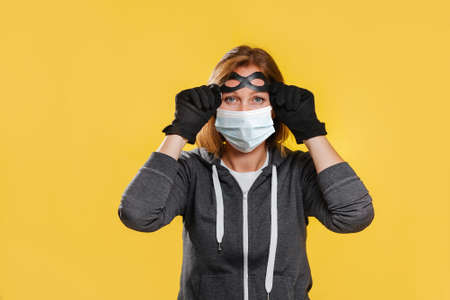 A female robber in a protective mask and gloves, puts on glasses. Yellow background. The concept of...