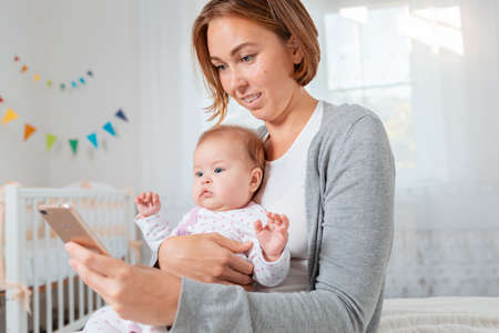 Portrait of a smiling mother holding a baby in her arms and using a smartphone. Childrens room on...