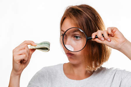 Portrait of a woman looks through a magnifying glass at a small wad of dollars. White background. The concept of checking counterfeit money.の写真素材