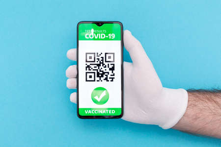 Concept of vaccination and digital passport. Hand in protective glove holding a cellphone with electronic Immunity passport with a COVID-19 vaccination stamp. Blue background. Flat lay.の写真素材