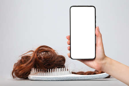 A white comb with false hair. White background. A female's hand holds a smartphone with a white screen. Mock up. The concept of buying and selling hair.の写真素材