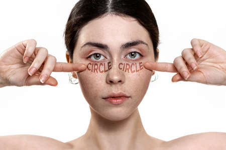 Close-up portrait of a young woman pointing at dark circles under her eyes. White background. Concept of skin care and combating the first signs of aging.の写真素材