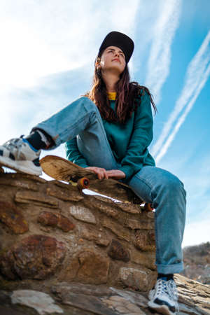 A young beautiful Caucasian woman sits on a skateboard and looks away. Bottom view. In the background, the blue sky with clouds. Vertical. Concept of sports lifestyle and street culture.の写真素材
