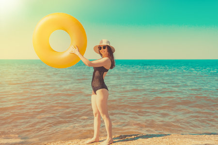 Young slim woman with glasses and hat is holding in her hands an orange circle swimming and enjoys the holiday. Sea on the background.The concept of holiday resort and travel. Tint and copy space.の写真素材