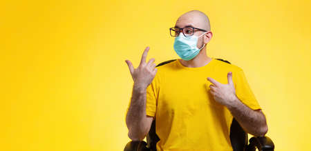 A bald man with glasses in medical mask sits in a chair and gestures emotionally. Yellow background. Copy space. Concept of self-isolation, quarantine, and virus protection.の写真素材