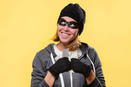 A female hacker in a black hat and a mask-glasses, greedily holds a wad of money in her hands and smiles. Yellow background. The concept of crime and theft.の写真素材