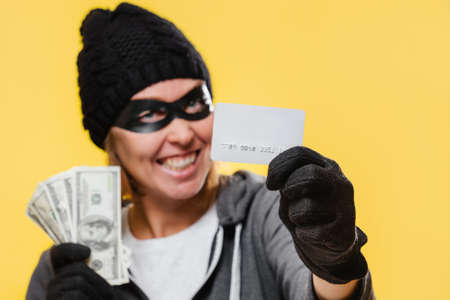 A female hacker wearing a black hat and a mask-glasses, smiling maliciously and holding a wad of money and a bank credit card. Yellow background. The concept of theft and cybercrime.の写真素材