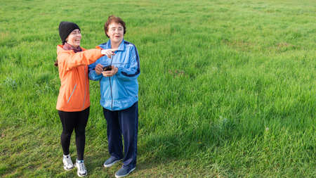 Happy adult granddaughter shows the direction to her grandmother with a smartphone in her hands. Grass in the background. Copy space. International Day of the Elderly.の写真素材