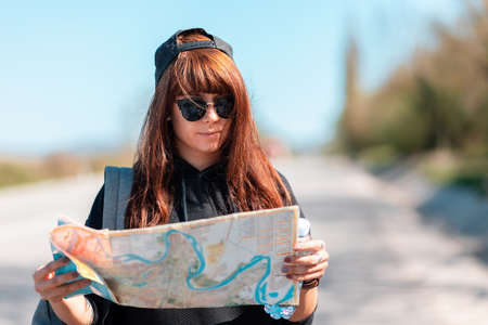 Local Travel. Portrait of young woman wearing a cap and sunglasses, holding a paper map. In the background, the road is blurred. The concept of hitchhiking.の写真素材